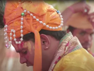 A man in profile wearing an orange head-dress, also draped with pearls. His brown is marked with red and yellow pigment.