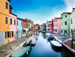 A canal in Italy with boats anchored alongside. There are colorful houses lining each side of the canal as we look down its length.