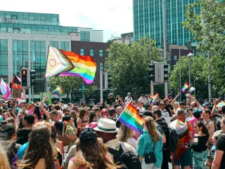 A group of people gathering in a public place. Several are flying rainbow Pride LGBTQIA+ flags, with a large Pride flag also featuring black and brown stripes, light blue, pink, and white stripes, and an Intersex solidary symbol.