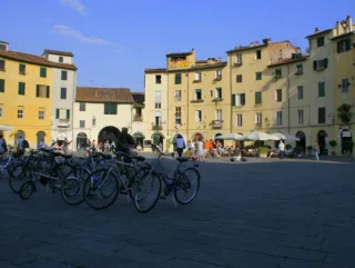 An Italian town plaza with parked bikes and people walking around. It is surrounded by houses.