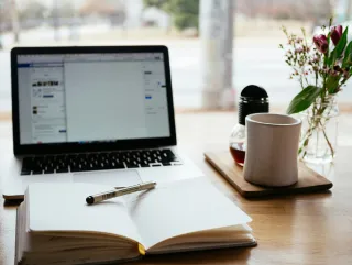 A stock photo of a desk with a laptop, coffee mug, notebook, and vase of flowers.