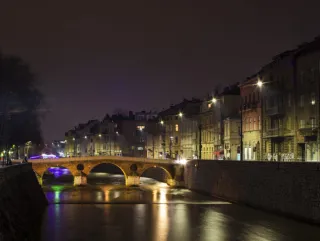 A city canal at night, spanned by a footbridge with three arches. The canal and houses are lit from below.