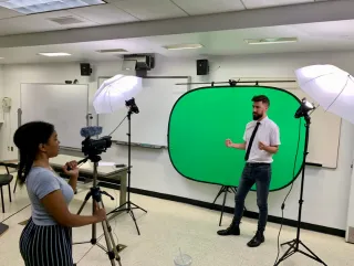 A man stands in front of a green screen. He is being recorded in a studio by a woman with a camera.