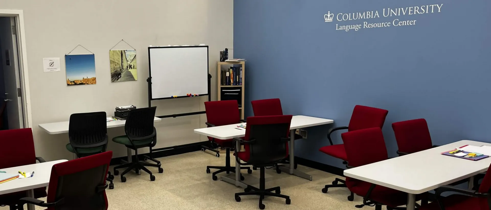 An image showing the interior of the Offline Writing Lab, with several tables and chairs, a small bookshelf, a whiteboard, and a blue wall with the logo of the Columbia Language Resource Center.