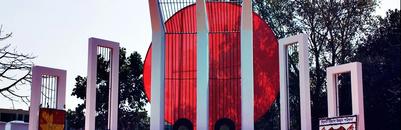 An image of the Shaheed Minar monument in Bangladesh, adorned with wreaths of orange flowers and Bengali banners in front of its white columns and large red circle.