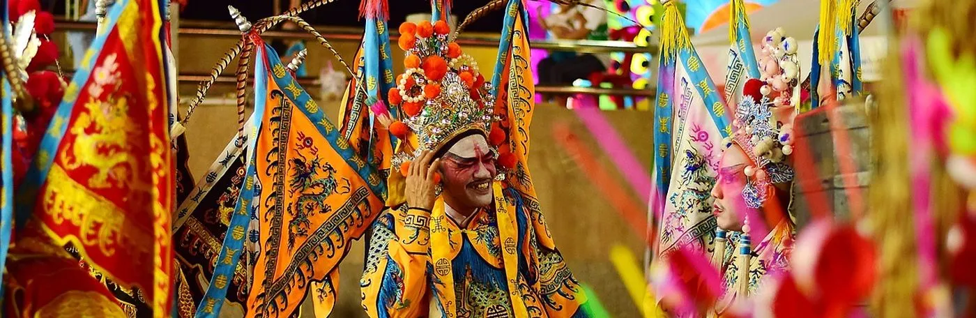 A vibrant close-up of Cantonese opera performers wearing elaborate, colorful costumes and ornate headdresses surrounded by decorative flags.