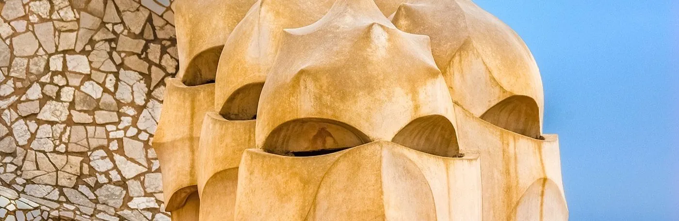 A close-up of Antoni Gaudí's sculptural chimneys on the rooftop of Casa Milà in Barcelona, set against a bright blue sky and a mosaic-patterned wall.