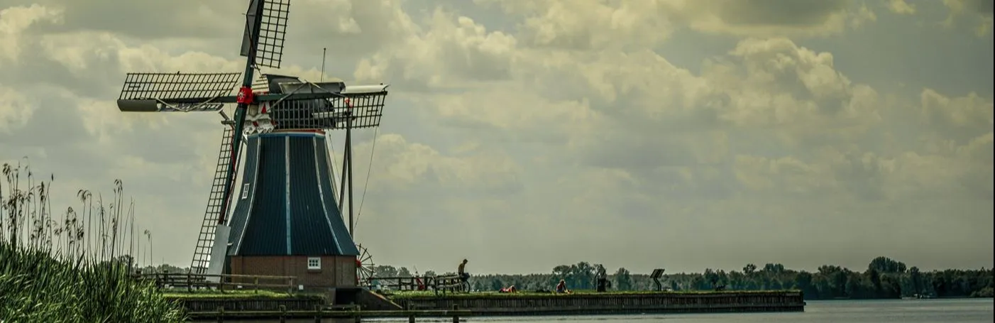 A large windmill with sails retracted and several people sunning on the shores of a river.
