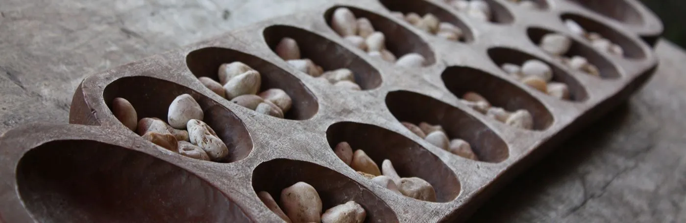 A magnified image of a handmade mancala board, hewn from wood and containing small stones as playing pieces.