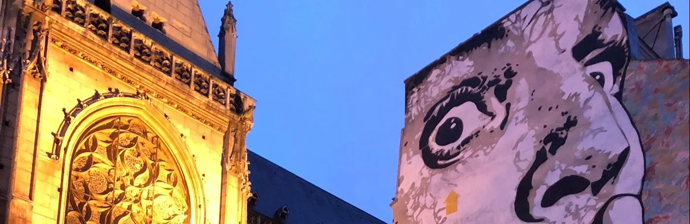 An illuminated gothic cathedrale faces a work of graffiti on an opposing building, featuring a wide-eyed man holding up his index finger to his lips in a shushing gesture.
