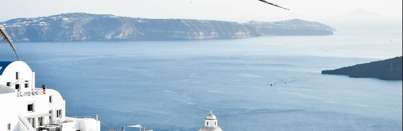 A scenic overlook of Santorini's white-washed buildings and domed structures nestled on the hillside, facing the Aegean Sea with volcanic islands in the background.