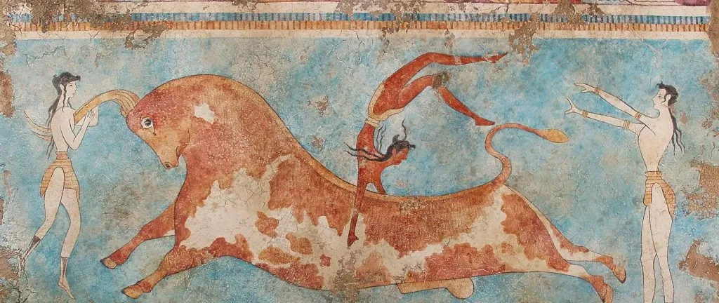 An ancient Minoan fresco depicting the bull-leaping ritual, with acrobatic figures performing stunts over a charging brown-and-white bull against a blue background.