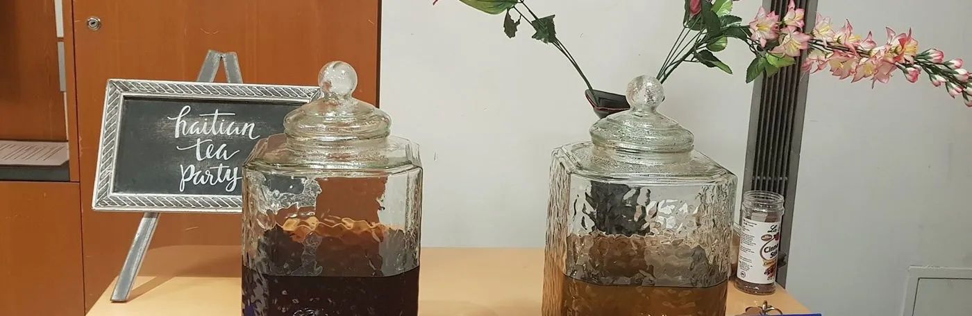 Two large glass beverage dispensers filled with tea sit on a table next to a small chalkboard sign that reads "haitian tea party," with flowers in the background.