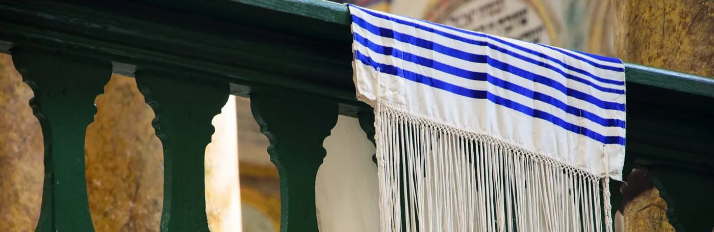 A white tallit (Jewish prayer shawl) with blue stripes and long, knotted fringes (tzitzit) draped over a dark green wooden railing inside a synagogue.