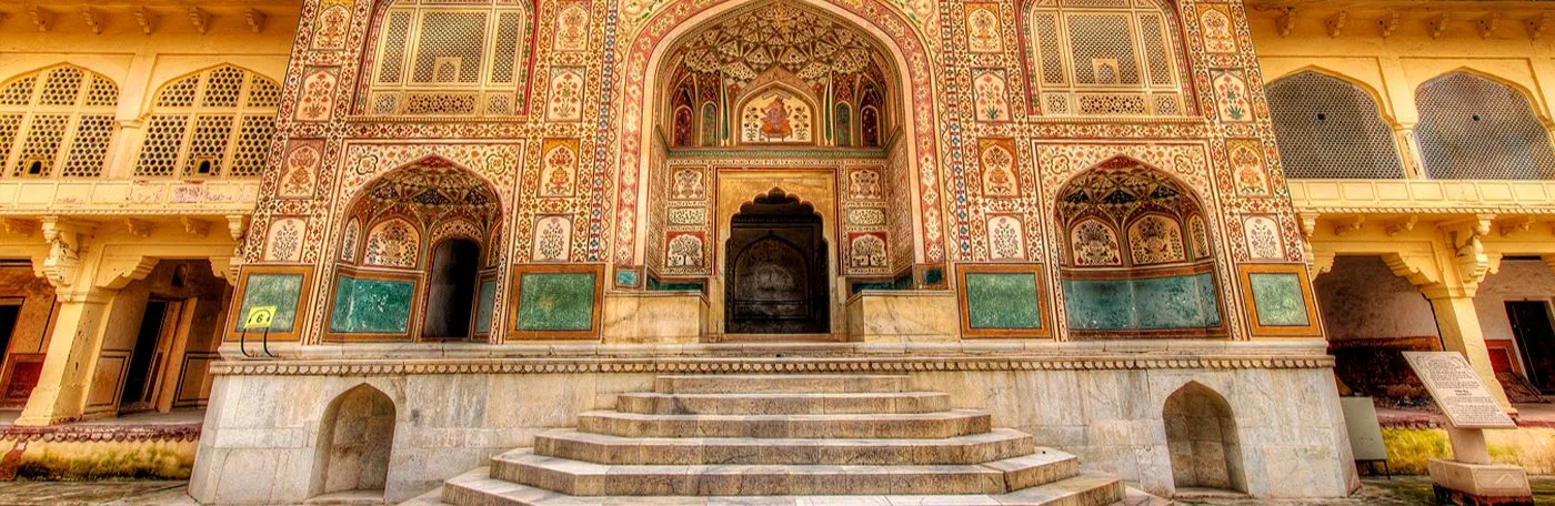 The ornate, multi-tiered entrance of Ganesh Pol at the Amber Palace in Jaipur, India, featuring detailed fresco paintings, mosaic work, and stone steps leading up to the main archway.