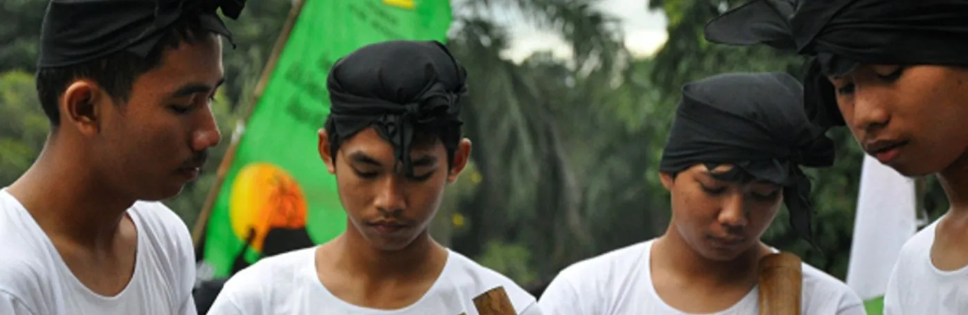 Four young men wearing white t-shirts and traditional black headbands look downward, with lush green trees and a colorful banner in the background.