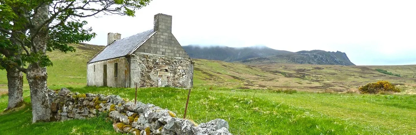 An old, stone cottage with a slate roof stands in a grassy field next to a low stone wall, with rolling green hills and misty mountains in the background.