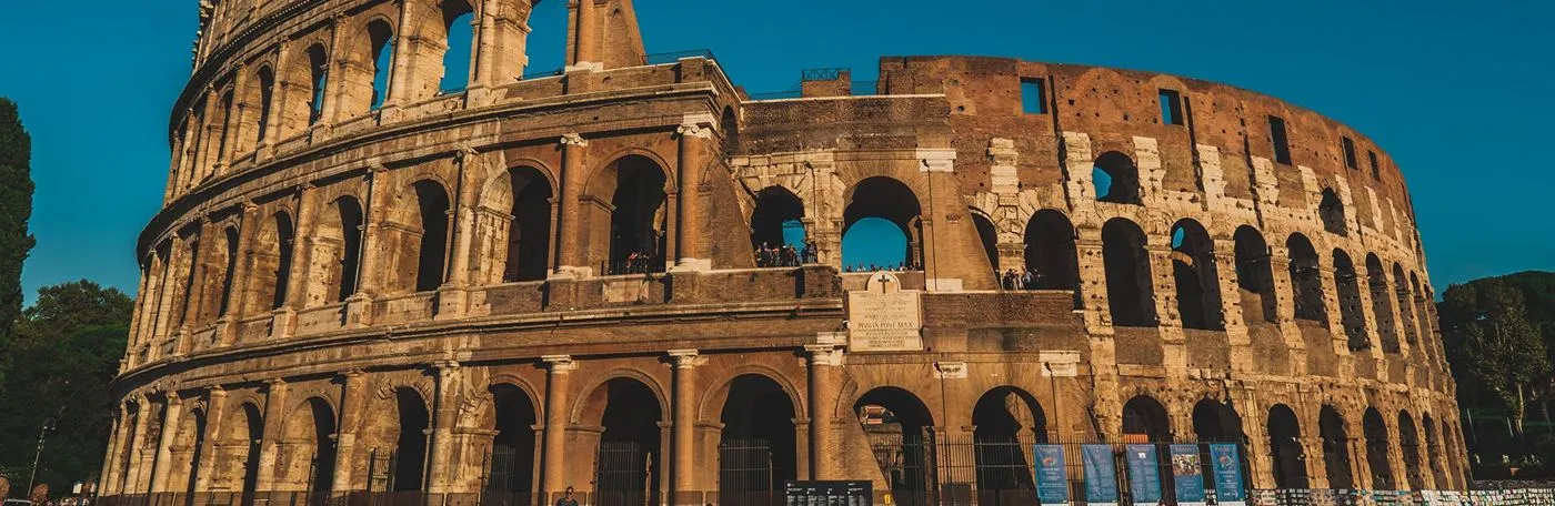 A wide-angle view of the ancient Colosseum in Rome, showing its iconic stone arches and tiered exterior walls under a clear blue sky.