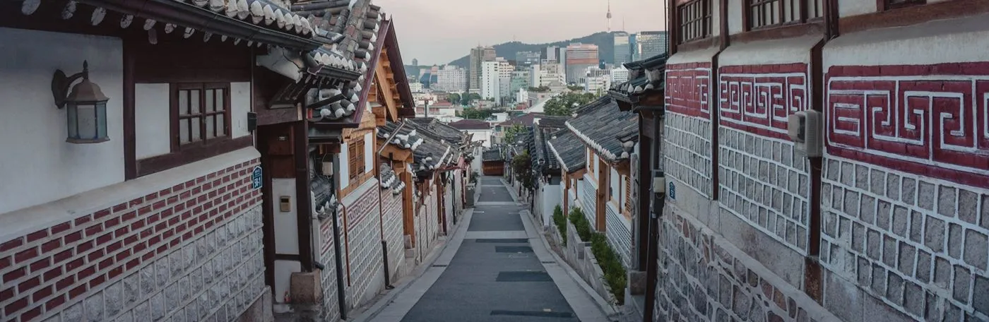 A view looking down a narrow, sloped street lined with traditional Korean hanok houses, with modern city skyscrapers and the N Seoul Tower visible in the distant background.
