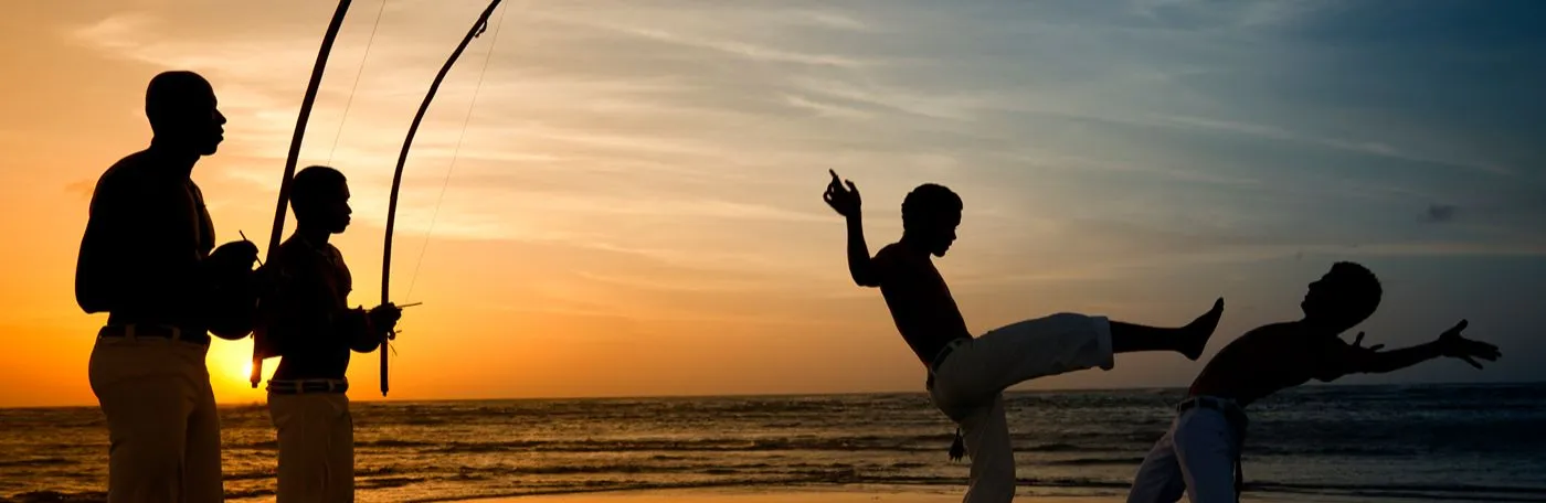 Two boys demonstrating Capoeira, the Brazilian martial art that combines elements of dance and music, in a beach scene at sunset.