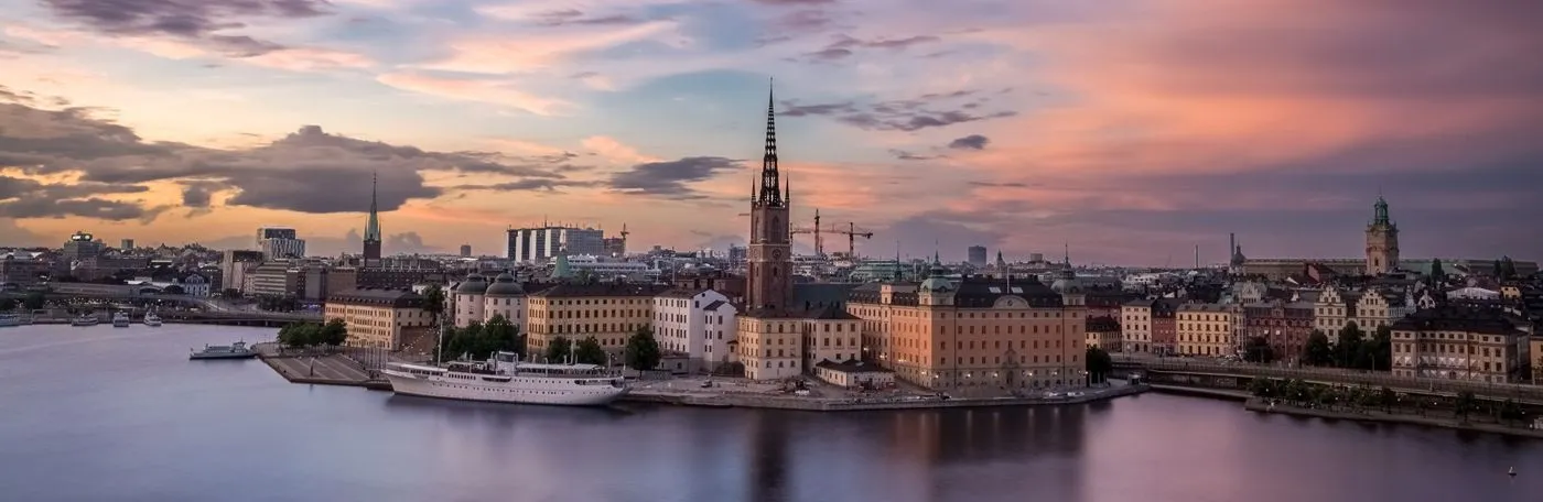 Panoramic twilight view of the Stockholm, Sweden waterfront and the Riddarholmen island skyline under a pink and orange sunset sky.