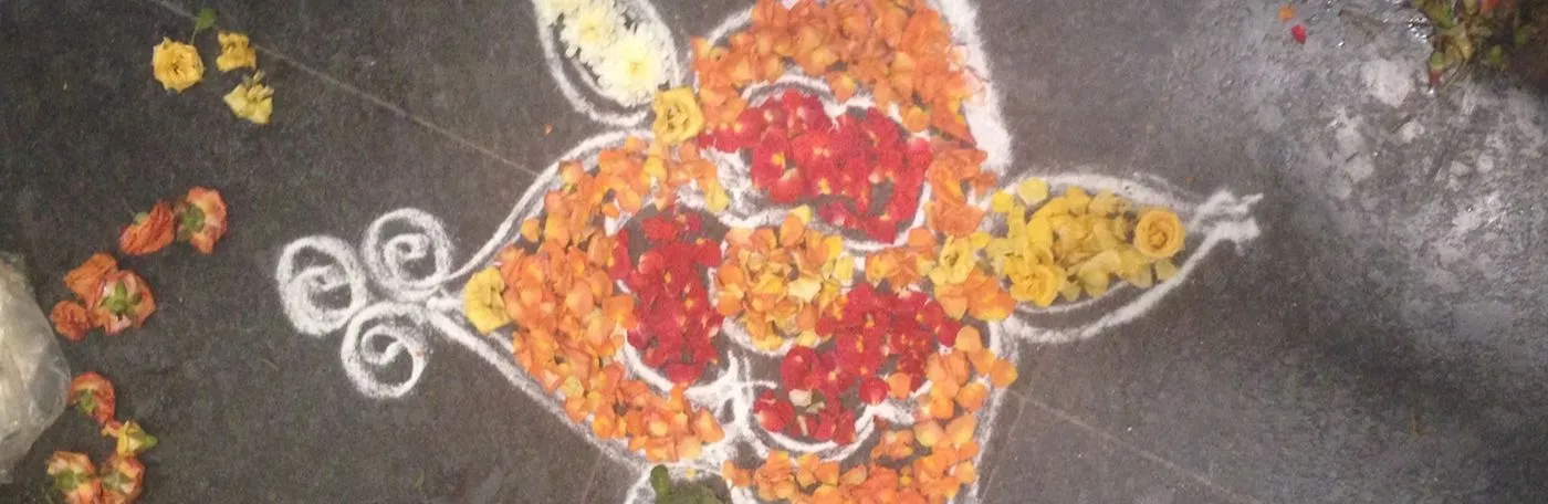 A traditional kolam or rangoli design outlined in white powder and filled with colorful flower petals on a dark floor.
