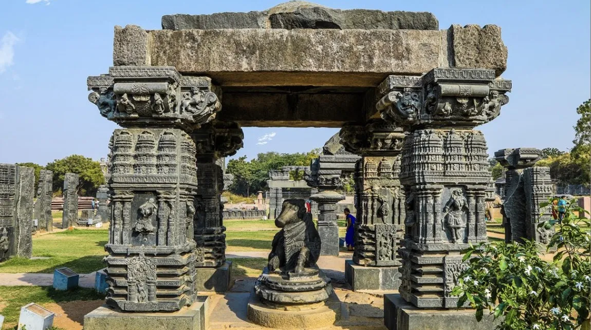 An ancient stone mandapa frame with intricately carved pillars housing a seated stone Nandi bull sculpture at the Warangal Fort ruins.