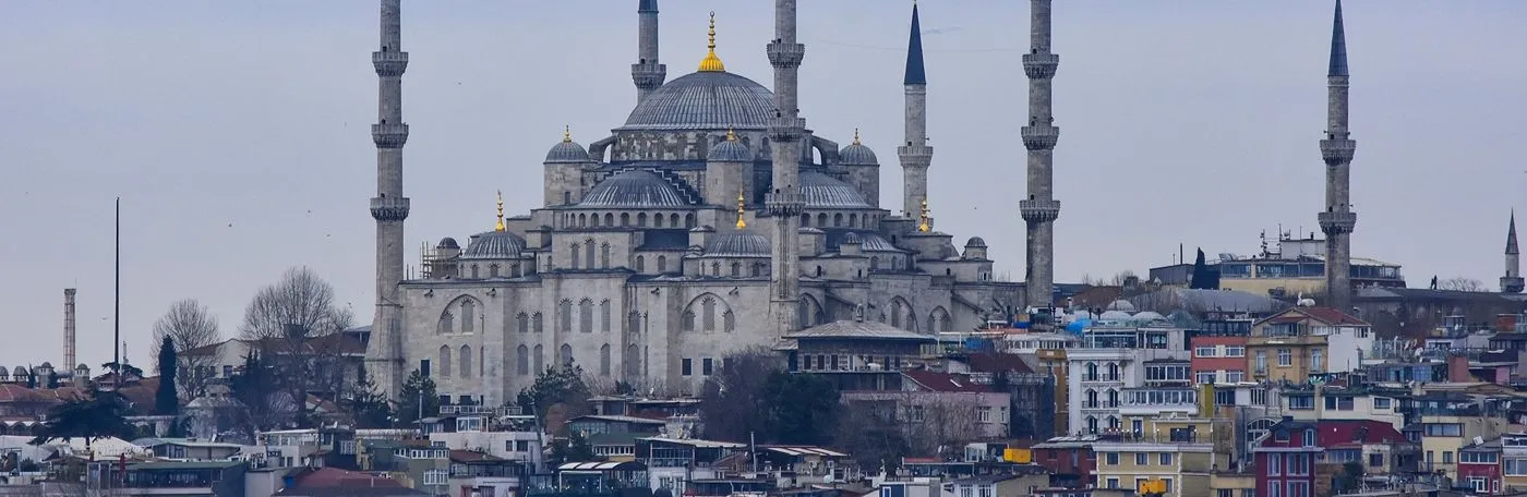 Panoramic view of the historic Sultan Ahmed Mosque (Blue Mosque) in Istanbul, Turkey, rising above the surrounding city buildings under a cloudy sky.