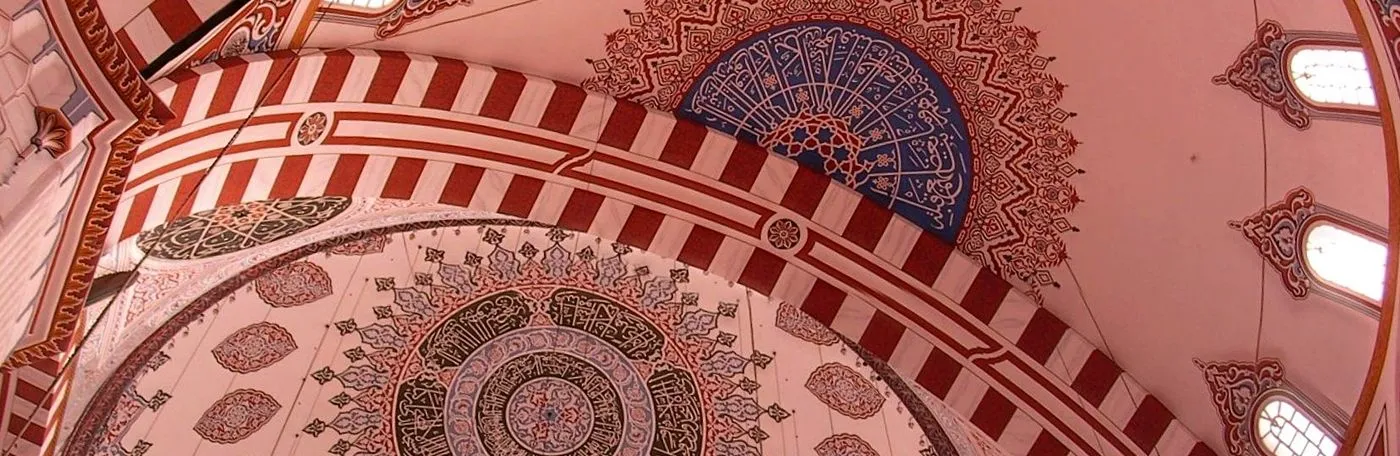 Low-angle panoramic view looking up at the ornately decorated domes and arches of an Ottoman-style mosque, featuring intricate Arabic calligraphy and red-and-white striped patterns.