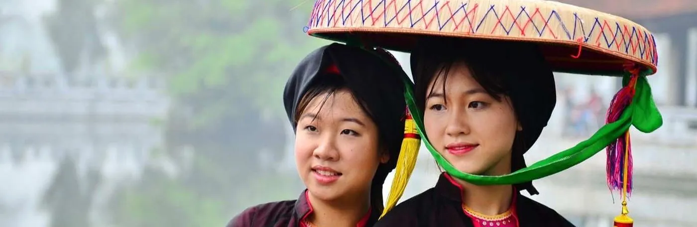 Two women wearing traditional Vietnamese garments and headwear standing outdoors against a blurred, foggy background.