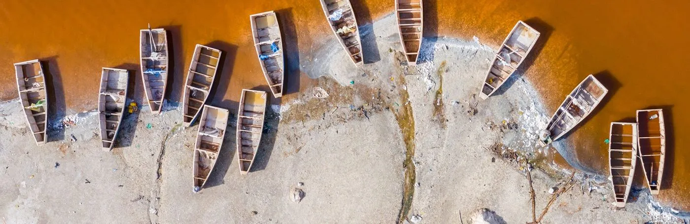 Top-down aerial photograph of traditional wooden boats lined up on the sandy shoreline of Lake Retba (Lac Rose) in Senegal, contrasting against the bright orange-pink water.