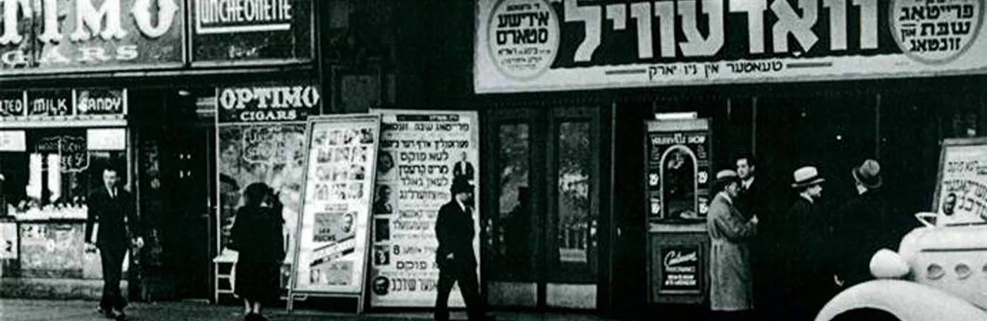 Historic black-and-white photo of a street scene in New York featuring a Yiddish theater storefront with signs and posters in Yiddish.