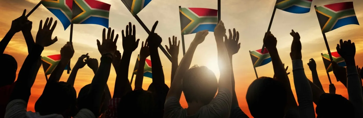 Silhouetted hands waving South African flags against a sunset sky.