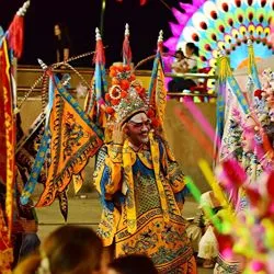 A vibrant close-up of Cantonese opera performers wearing elaborate, colorful costumes and ornate headdresses surrounded by decorative flags.