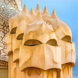 A close-up of Antoni Gaudí's sculptural chimneys on the rooftop of Casa Milà in Barcelona, set against a bright blue sky and a mosaic-patterned wall.