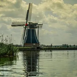 A large windmill with sails retracted and several people sunning on the shores of a river.