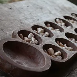 A magnified image of a handmade mancala board, hewn from wood and containing small stones as playing pieces.