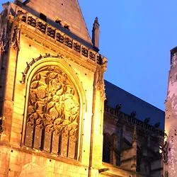 An illuminated gothic cathedral faces a work of graffiti on an opposing building, featuring a wide-eyed man holding up his index finger to his lips in a shushing gesture.