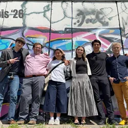 A group of six young adults poses together outdoors in front of a colorful graffiti wall.