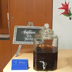 Two large glass beverage dispensers filled with tea sit on a table next to a small chalkboard sign that reads "haitian tea party," with flowers in the background.