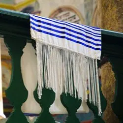A white tallit (Jewish prayer shawl) with blue stripes and long, knotted fringes (tzitzit) draped over a dark green wooden railing inside a synagogue.