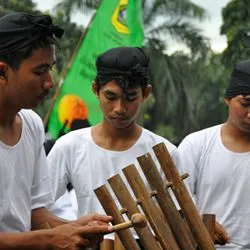 Four young men wearing white t-shirts and traditional black headbands look downward, with lush green trees and a colorful banner in the background.