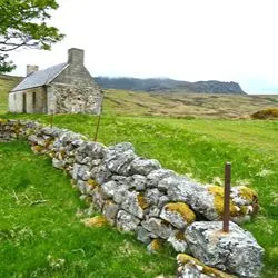 An old, stone cottage with a slate roof stands in a grassy field next to a low stone wall, with rolling green hills and misty mountains in the background.