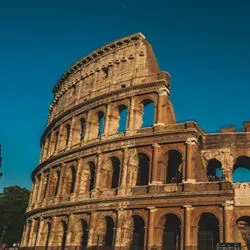 A wide-angle view of the ancient Colosseum in Rome, showing its iconic stone arches and tiered exterior walls under a clear blue sky.
