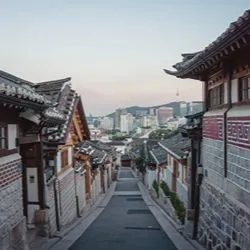 A view looking down a narrow, sloped street lined with traditional Korean hanok houses, with modern city skyscrapers and the N Seoul Tower visible in the distant background.