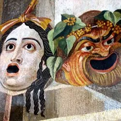 A Roman mosaic depicting two theatrical masks: a pale, wide-eyed tragedy mask on the left and a grinning, bearded comedy mask adorned with ivy leaves on the right.