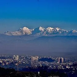 A panoramic view of a snow-capped mountain range rising above hazy, dark blue foothills under a clear blue sky.