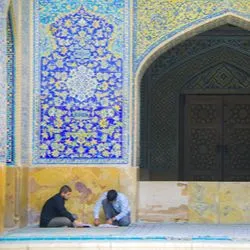 Two young men sit on rugs in an outdoor courtyard of a building adorned with intricate blue and yellow floral mosaic tiles, focusing on open books in front of them.
