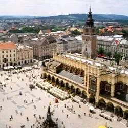 An aerial view of Krakow's Main Market Square in Poland, showing the historic Cloth Hall, St. Adalbert's Church, and pedestrians walking across the sunny plaza.