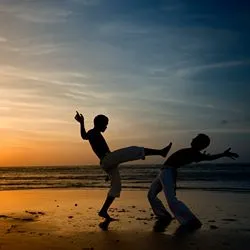 Two boys demonstrating Capoeira, the Brazilian martial art that combines elements of dance and music, in a beach scene at sunset.